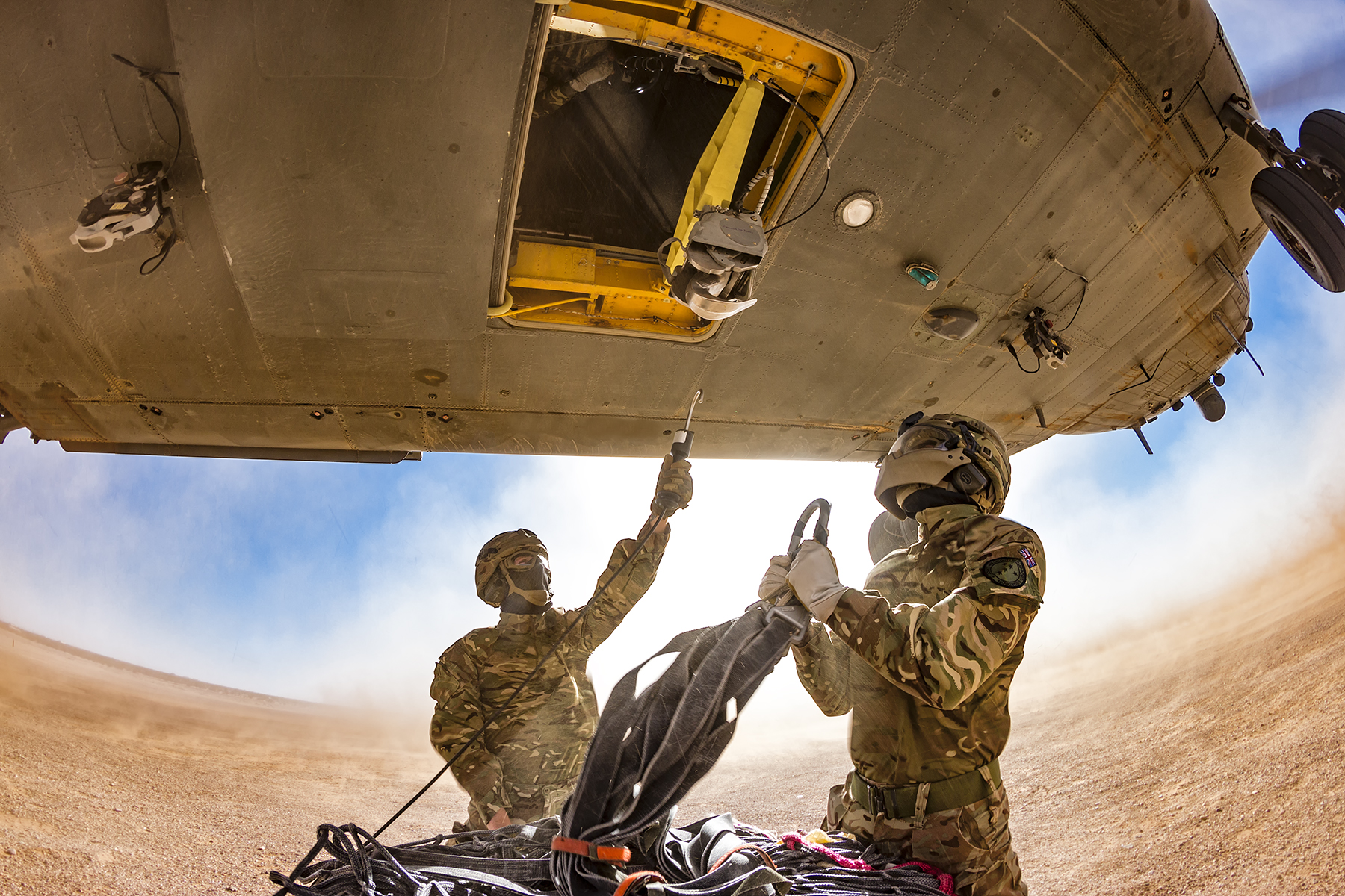 Image shows aviators attaching a load to the under-sling loads of a helicopter.